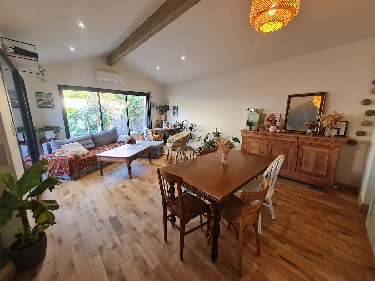 A spacious living area features large glass doors that provide natural light and views of the garden. A comfortable sofa is positioned near a coffee table, complemented by a dining table with various chairs. Warm wooden floors extend throughout the room.
