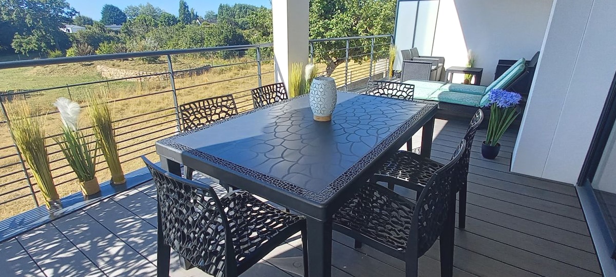 A spacious terrace features a black dining table surrounded by six black chairs. A decorative vase is placed at the center of the table. Lounge seating is visible in the background, with greenery and an open view of the surrounding landscape.