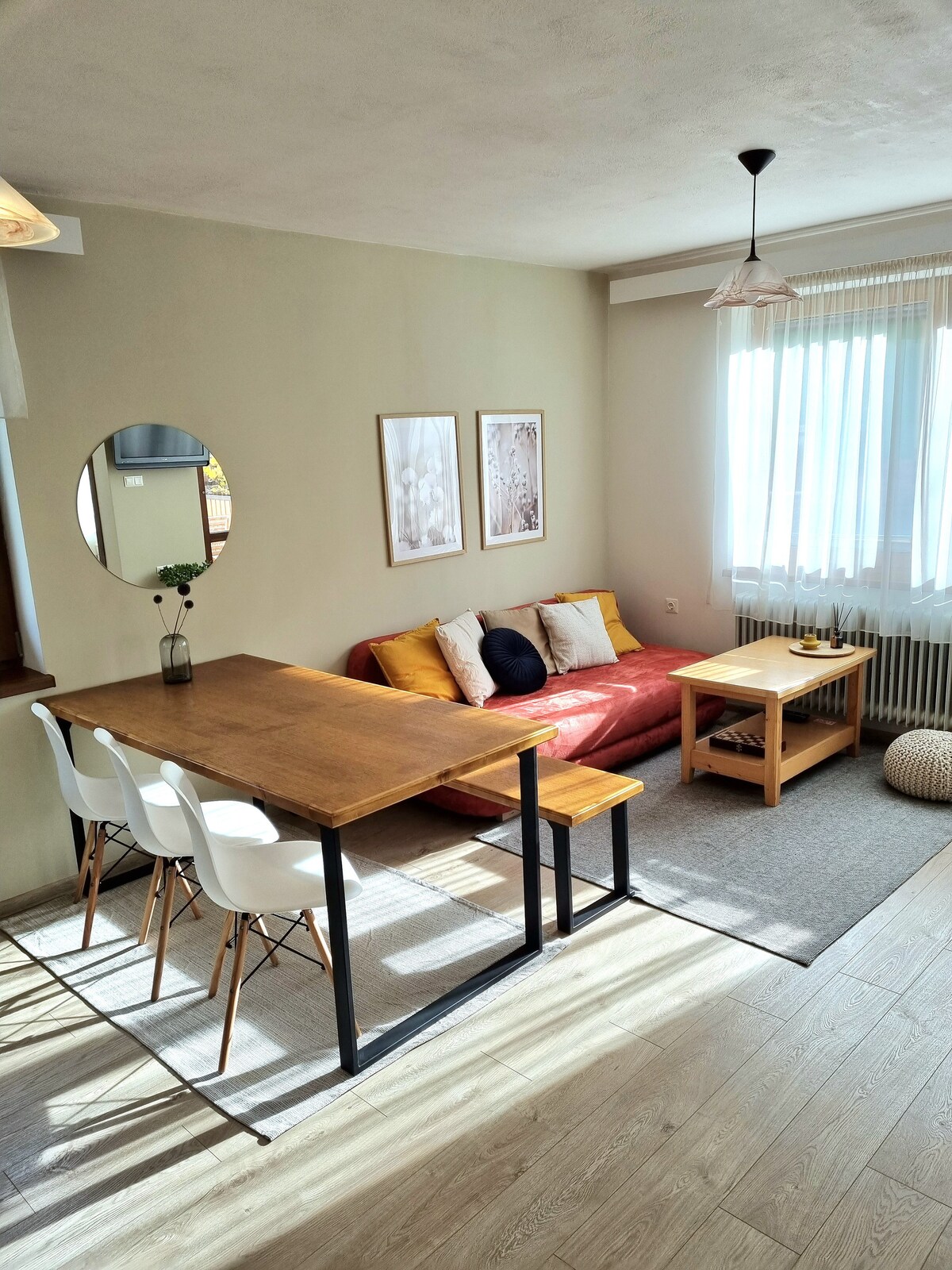 A bright living space features a red sofa adorned with cushions and two framed artworks on the wall. A wooden table is paired with white chairs, while sunlight filters through sheer curtains, creating a warm ambiance.