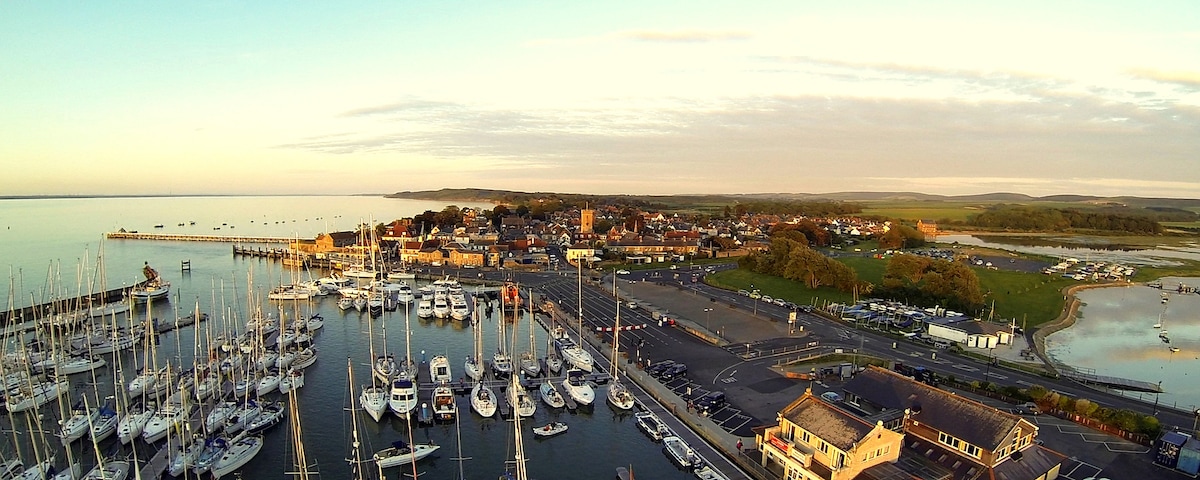 An aerial view captures the bustling marina filled with numerous sailboats and yachts. The scenery includes the vibrant town of Yarmouth in the background, with its buildings and green areas extending towards the coastline, showcasing a calm sea under a soft sunrise light.