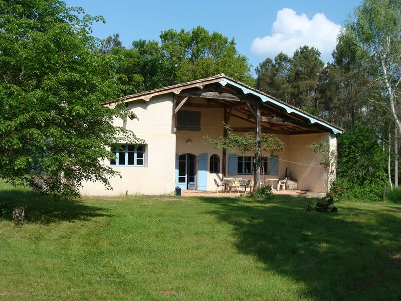 A spacious farmhouse is surrounded by greenery, featuring a large covered terrace with outdoor seating. Light blue shutters frame the windows, and the building's earthy tones blend with the natural landscape. The open area in front is bordered by trees and offers a serene atmosphere.