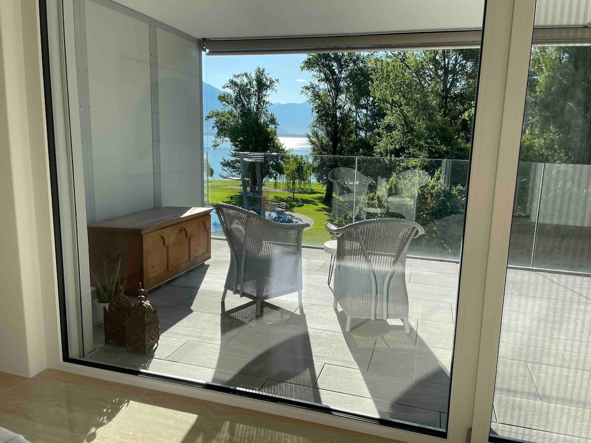 A view from inside reveals a spacious terrace featuring two comfortable chairs and a wooden storage chest. The terrace overlooks a scenic landscape with lush greenery and a glimpse of the lake in the distance, framed by large glass doors that invite natural light.