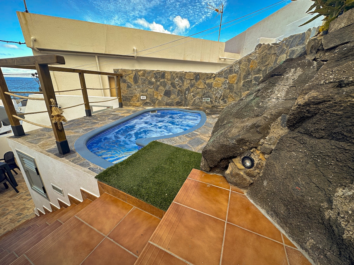 A secluded terrace features a private jacuzzi surrounded by natural stone and greenery. Steps lead down from the elevated area, emphasizing the integration of outdoor relaxation with the scenic surroundings. The distant view includes a clear sky and water, enhancing the tranquil setting.