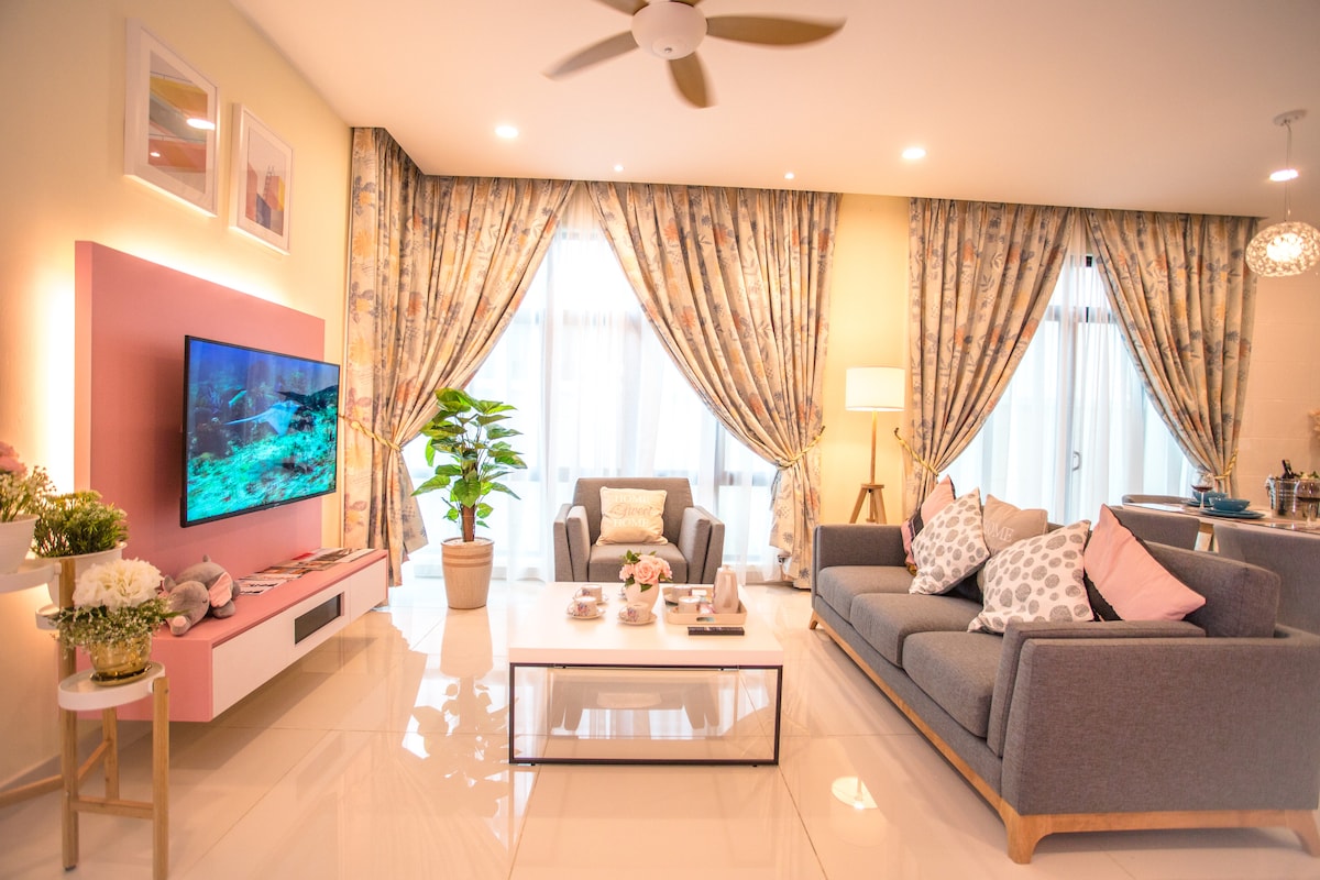 The living room features a spacious layout with a gray sofa adorned with decorative pillows, complementing a modern coffee table. A wall-mounted TV is positioned above a stylish pink entertainment unit. Large windows are dressed with patterned curtains, allowing natural light into the room.