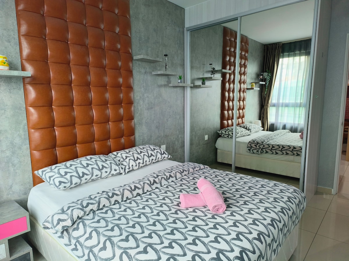 A stylish bedroom features a large bed with a decorative heart-patterned duvet cover. The headboard presents a tufted brown design. Mirrored wardrobes reflect the room's space, while soft natural light enters through sheer curtains, enhancing the modern ambiance.