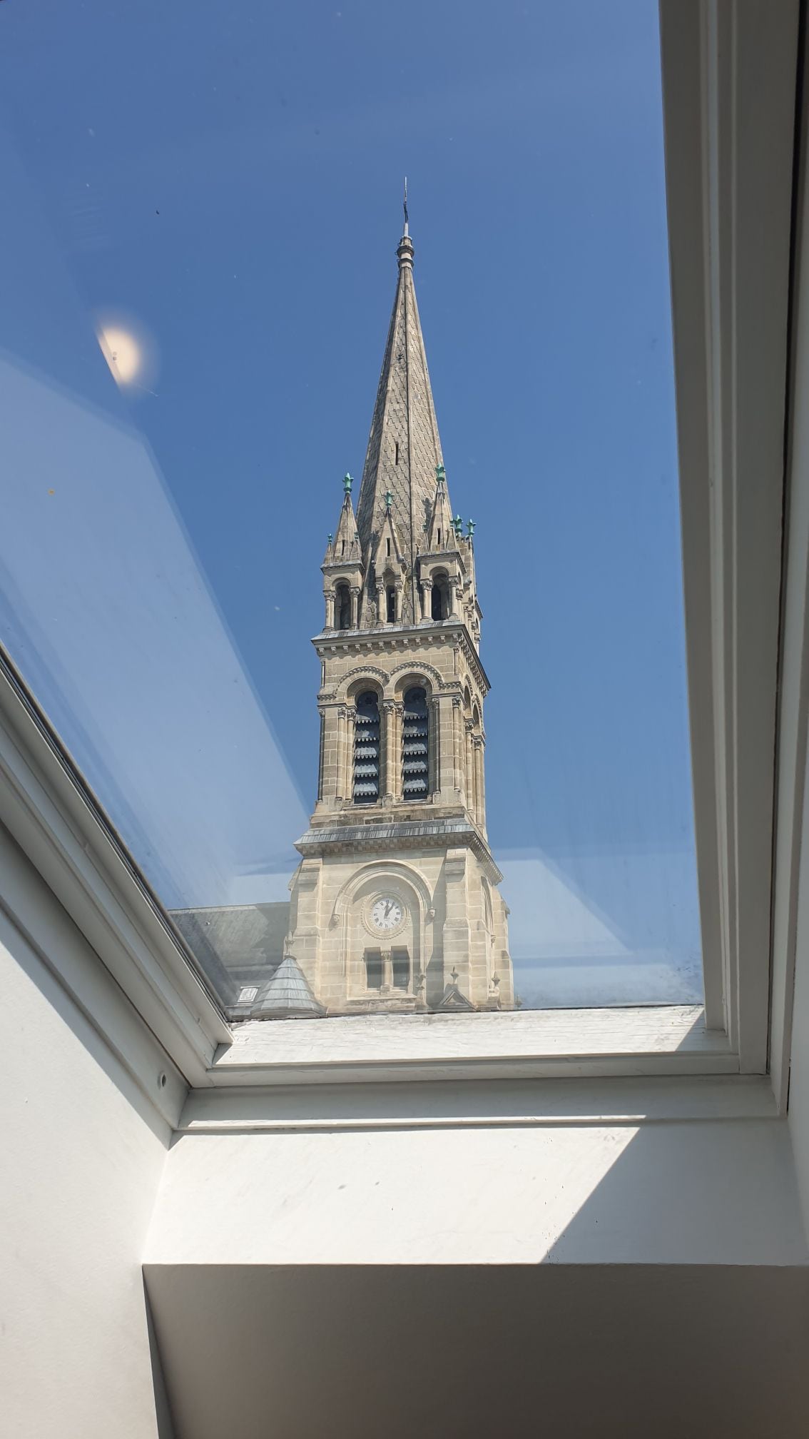 A view of a tall church steeple can be seen through a skylight. The steeple features intricate architectural details, with a clear blue sky in the background, enhancing the serene setting.