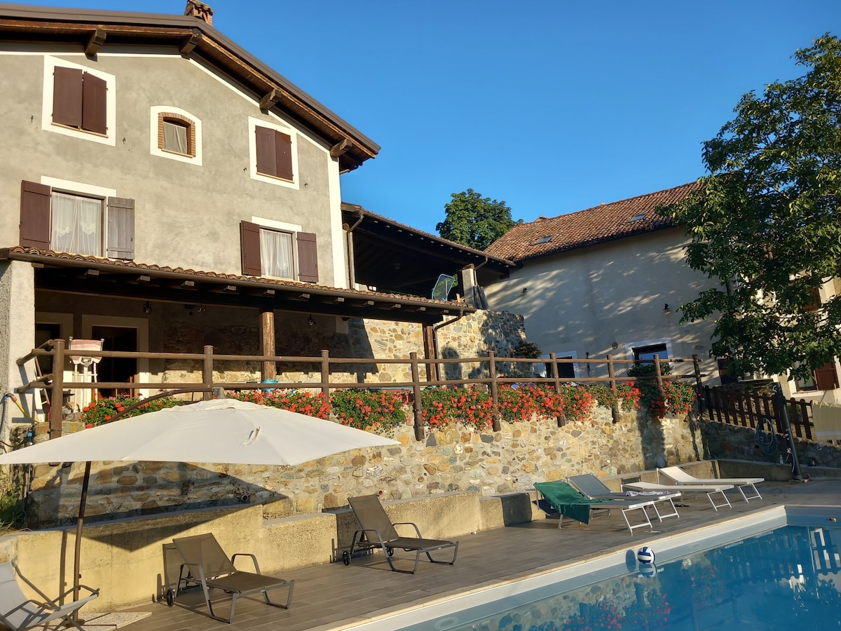 A two-story house with a rustic stone facade is complemented by wooden shutters and a spacious terrace. Lounge chairs surround the inviting swimming pool, while colorful flowers enhance the outdoor space. A shaded area with umbrellas is available for relaxation by the poolside.