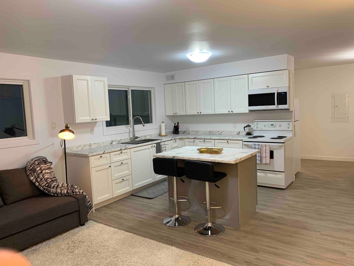 A modern kitchen layout is presented, featuring white cabinetry and a spacious island with bar seating. Light from the overhead fixture illuminates the room, highlighting the marble countertops. Windows allow natural light, and appliances include a microwave and stove.