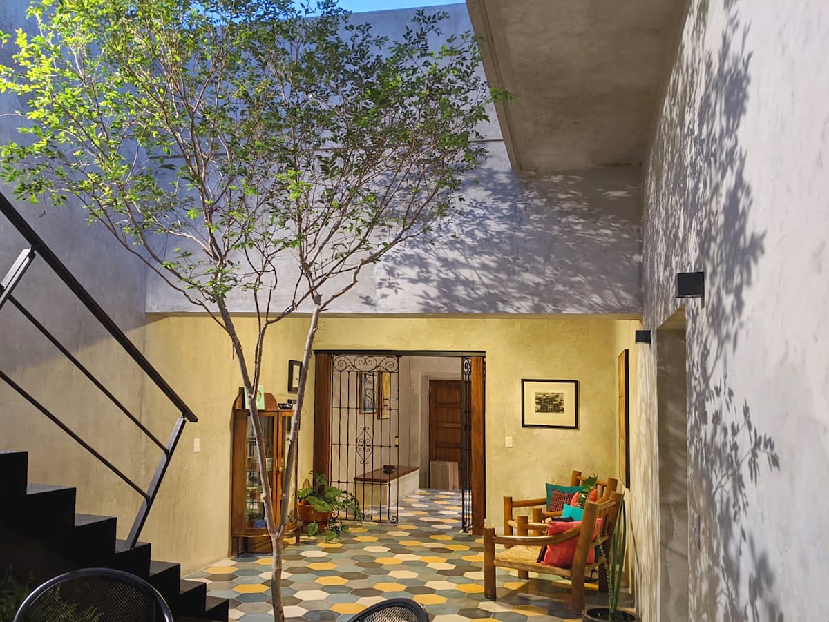 A spacious interior area is illuminated by natural light, featuring a central tree that adds a touch of greenery. The floor is accented with colorful tiles, while wooden furniture and a decorative railing enhance the welcoming atmosphere.
