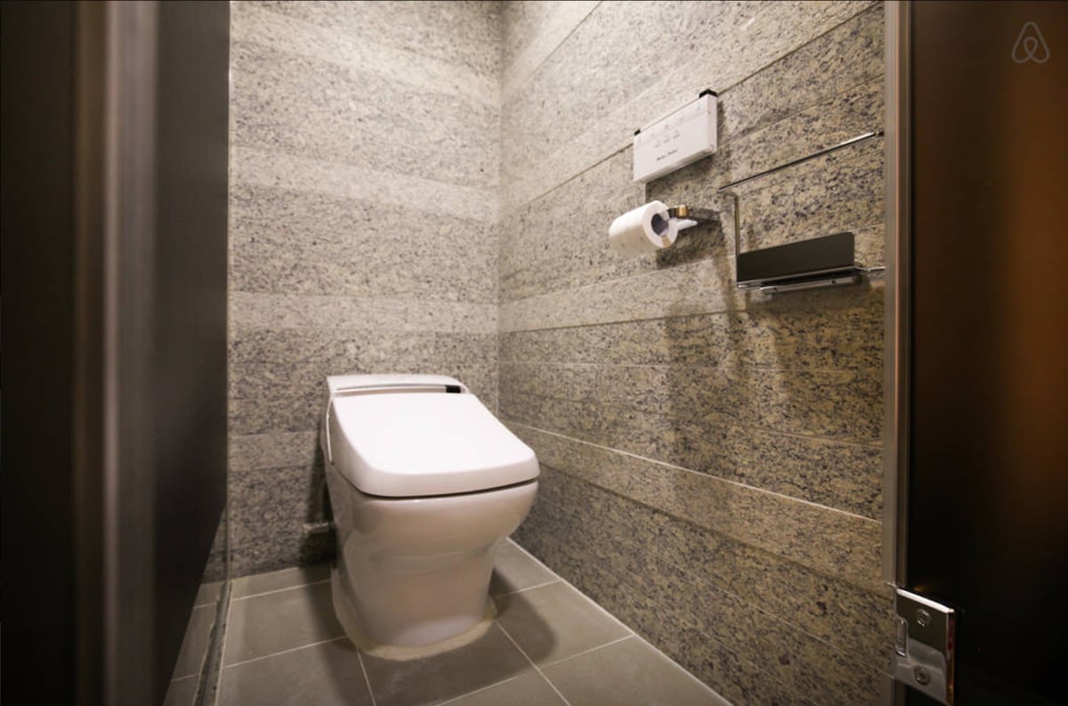 A well-appointed bathroom features a modern toilet against textured stone walls. Soft lighting enhances the clean lines of the space, while a roll of toilet paper and a toilet paper holder are conveniently positioned nearby.