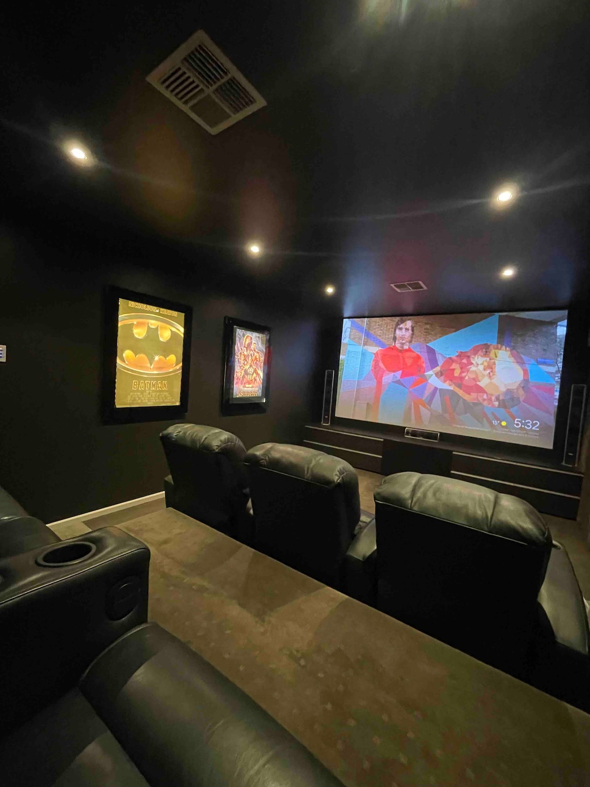A home theatre room features dark walls, equipped with a large screen displaying a colorful image. Plush black seating accommodates multiple guests, with built-in cup holders. Movie posters adorn the walls, adding to the cinematic atmosphere.