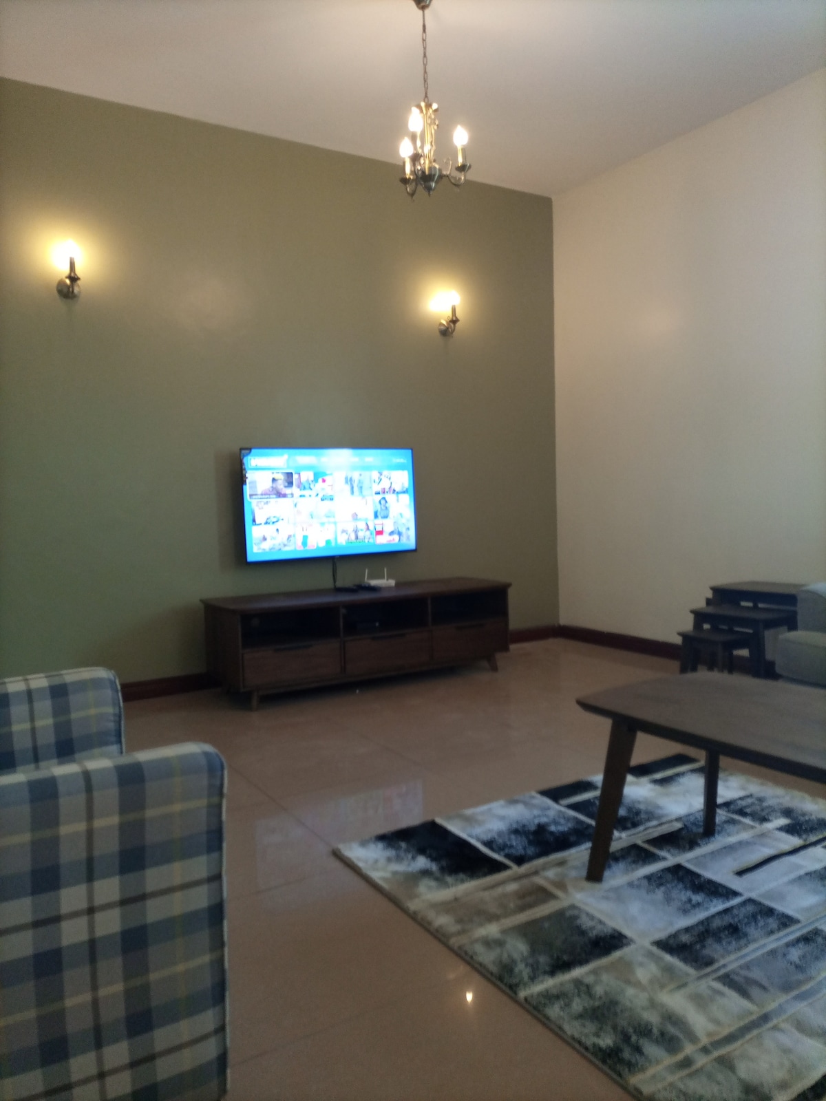 A comfortable living area features a large flatscreen television mounted on the wall across from two cozy armchairs. A wooden coffee table sits on an area rug, with soft lighting provided by wall sconces and a chandelier above.