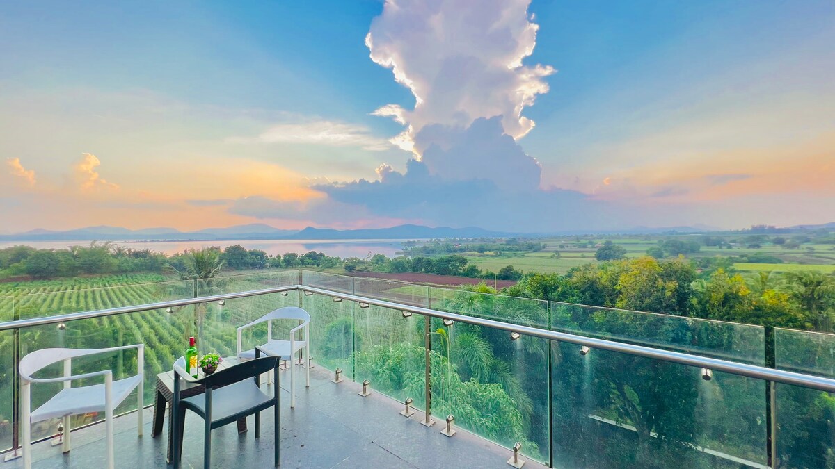 A serene balcony offers stunning views of lush green fields and the backwaters of Gangapur Dam. Two white chairs and a small table are positioned on the glass railing, with the colorful sky reflecting the sunset in the background.