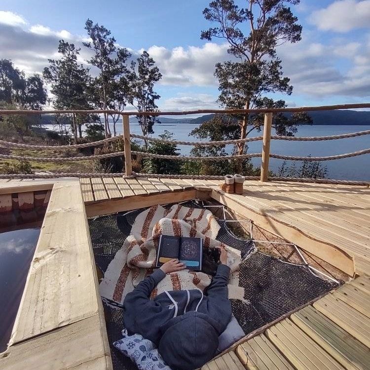 A person relaxes on a cozy blanket in a suspended net area overlooking a serene body of water. The surrounding landscape includes green trees and cloudy skies, providing a calm and inviting setting for relaxation.