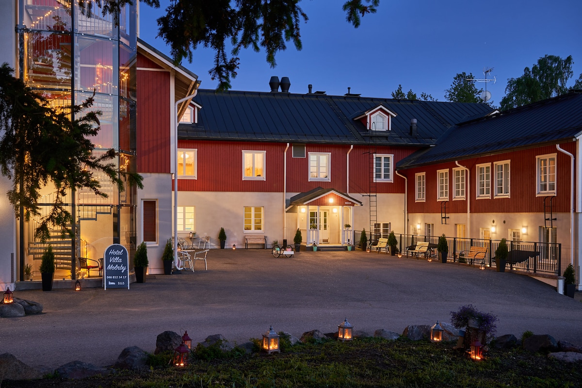 The hotel exterior is highlighted by charming red and white facades, framed by lush greenery. Soft lighting from lanterns creates a welcoming ambiance, while the entrance features an inviting pathway leading to the warmly lit entrance. A glass tower is visible, adding to the unique architectural design.