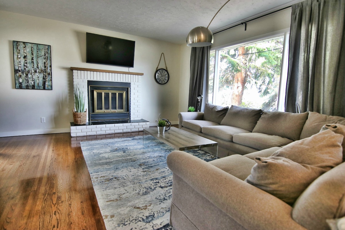 A cozy living area features a large sectional sofa arranged around a glass coffee table. A decorative fireplace is framed by white bricks, while a flat-screen TV is mounted above. Natural light enters through the window, revealing views of nearby trees.