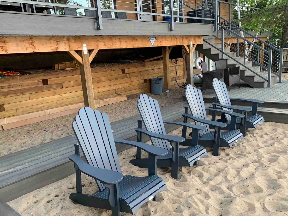 Four adirondack chairs are arranged neatly in a sandy area near the cabin, facing towards the lake. The chairs are a dark color with a slatted design. The wooden deck of the cabin is visible in the background, along with a staircase leading to the entrance.