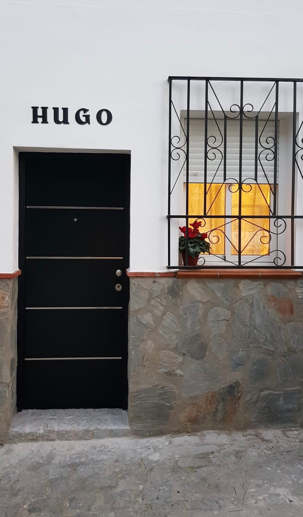 The entrance features a black door with the name 'HUGO' displayed above. A decorative window is adorned with a flower pot, providing a touch of color. The stone wall adds a rustic element to the inviting façade.
