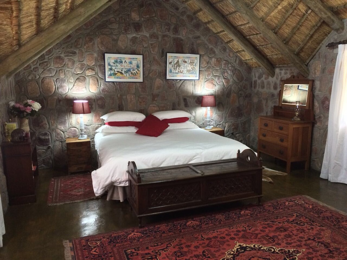 A cozy sleeping area features a spacious bed adorned with crisp white linens and red accent pillows. Stone walls provide a rustic backdrop, complemented by wooden nightstands with lamps. A patterned rug adds warmth to the floor, while framed artwork enhances the inviting atmosphere.
