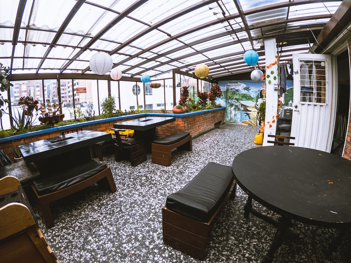 A covered outdoor terrace area features several dark wooden tables with integrated seating. Colorful paper lanterns hang above, and lush greenery is positioned along the edges. The floor is adorned with light-colored decorative stones, creating a unique ambiance for guests to enjoy.