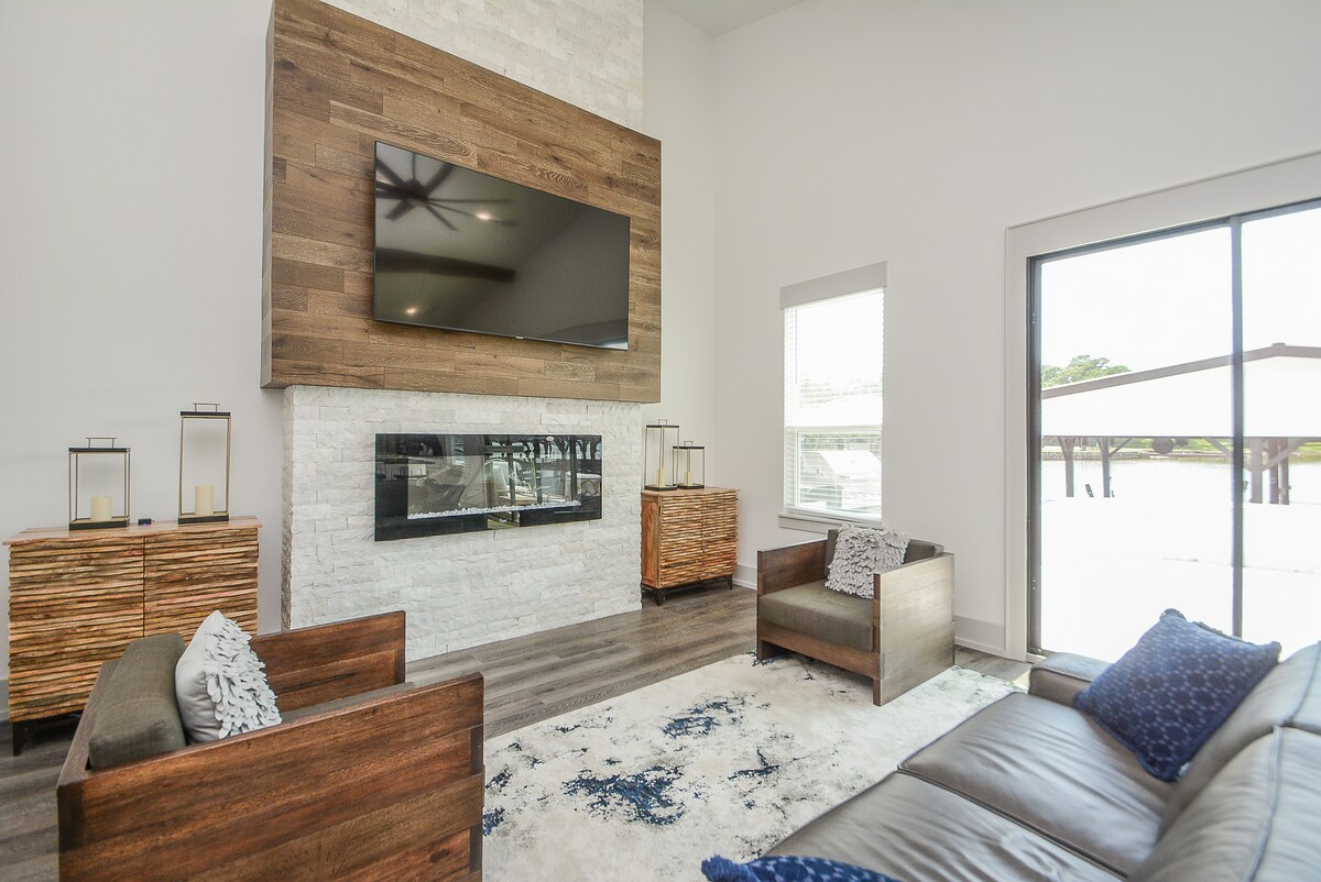 A spacious living area features a modern design with a large wall-mounted TV above a linear fireplace. Two armchairs and a sectional sofa surround a textured area rug, while large windows offer views of the outdoor space. Light-colored walls complement the wood accents.