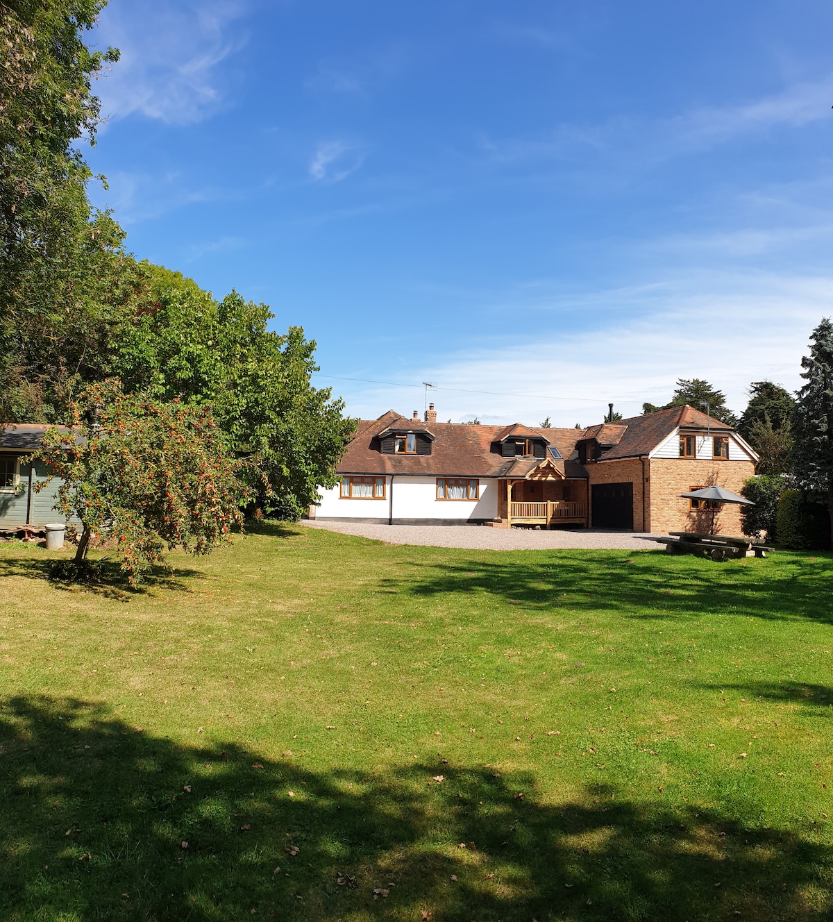 A well-kept garden surrounds the cottage, featuring a spacious lawn with a variety of trees. The cottage's façade includes multiple windows and distinct architectural features, while the driveway leads to the parking area beside the building.
