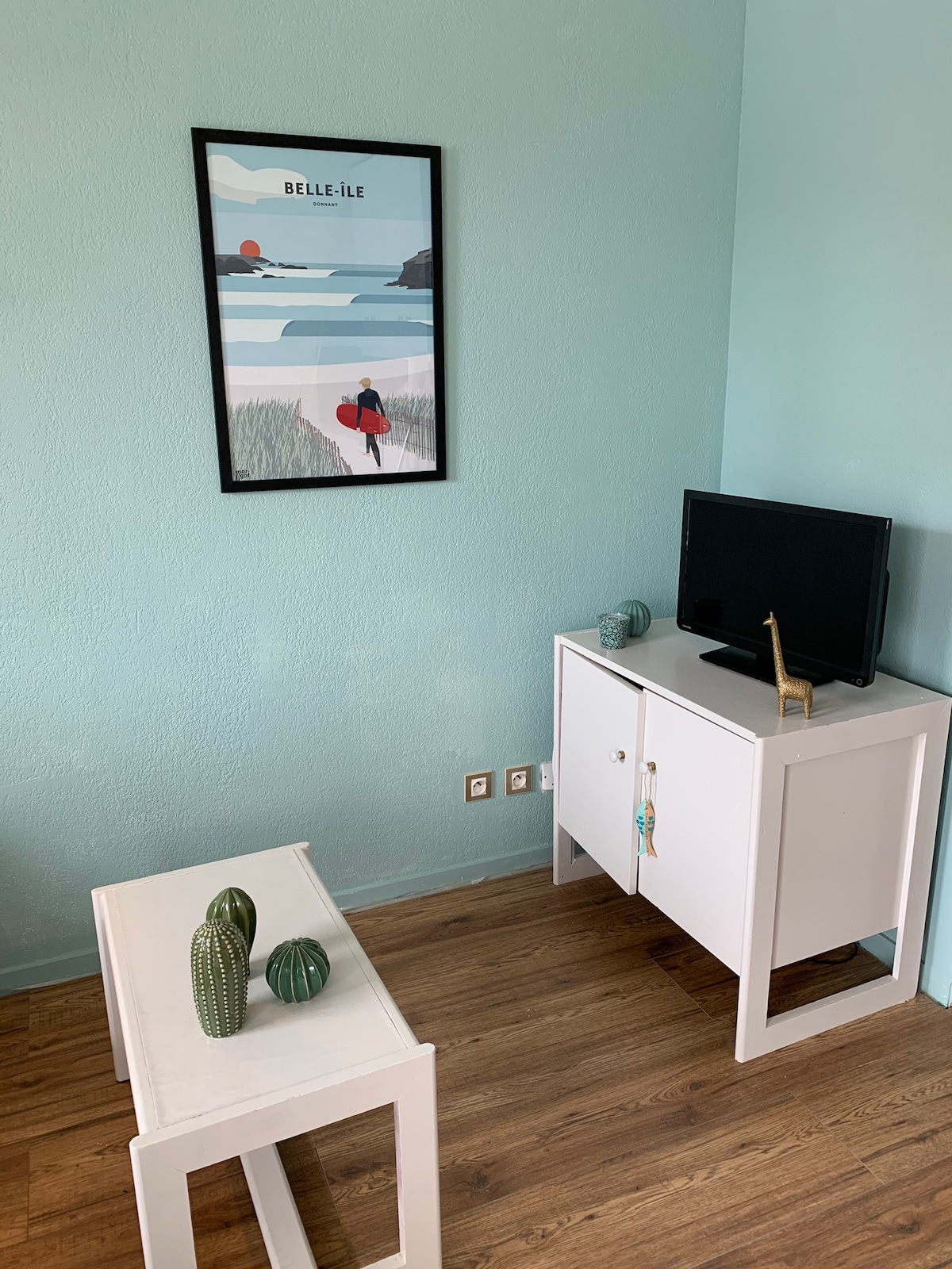 A light blue wall creates a serene backdrop for a framed artwork depicting a landscape with a sunset. A white cabinet with a TV on top is positioned beside two small white tables, adorned with green decorative items resembling cacti.