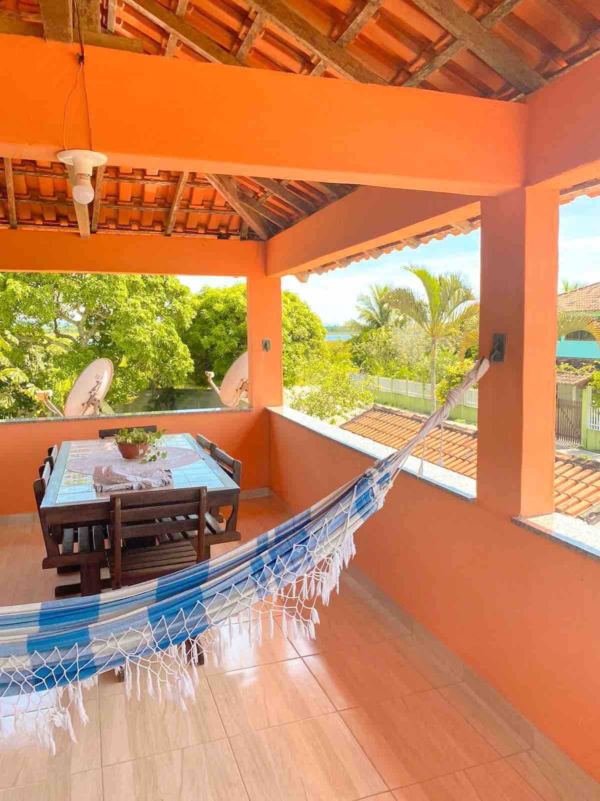 A bright balcony area features an inviting wooden dining table set for four, surrounded by lush greenery. A hammock hangs nearby, providing a relaxing spot to enjoy the view of the tranquil surroundings. Sunlight filters through, highlighting the vibrant orange walls.