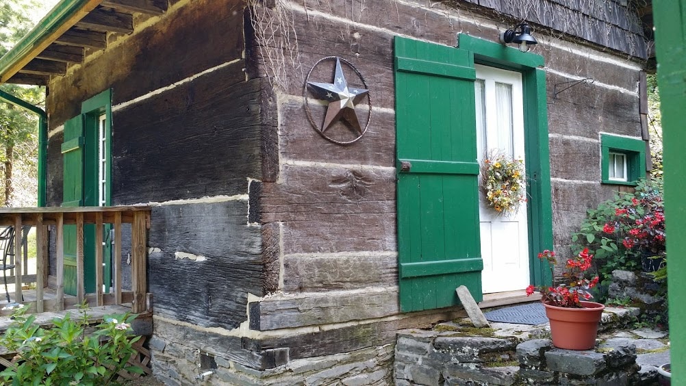 The exterior of a log cottage is prominently displayed, featuring rustic wooden logs and vibrant green trim. A white door adorned with a wreath is visible, accompanied by a star decoration on the wall. Flower pots with red blooms contribute to the welcoming appearance.