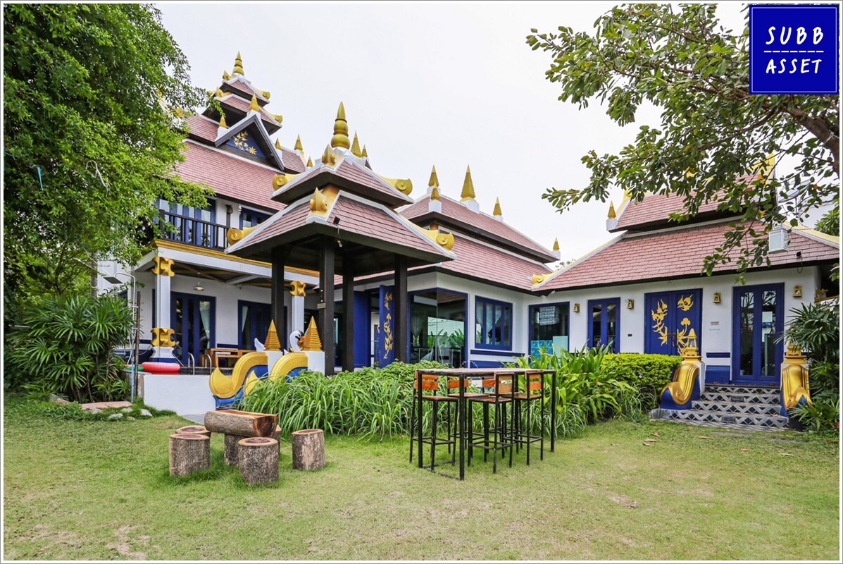 A traditional Thai-style villa is surrounded by lush greenery, featuring ornate golden rooftops and vibrant blue accents. Outdoor seating is provided at a small table alongside wooden stools, complemented by landscaped grass.