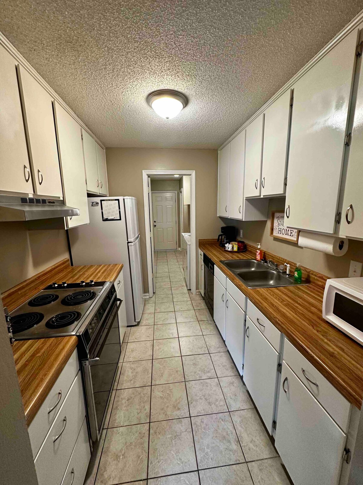 The kitchen features a narrow layout with white cabinetry and light wood countertops. An electric stove and oven are positioned alongside a stainless steel sink. A refrigerator and microwave are included, with tile flooring that extends toward a hallway in the background.