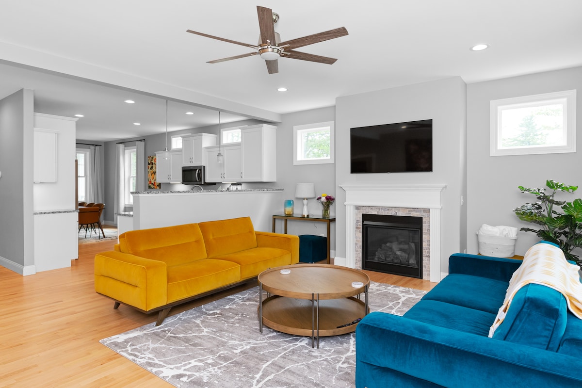 The living room features two sofas in vibrant colors, one in mustard yellow and the other in blue, arranged around a round coffee table. A gas fireplace is centered along the back wall, beneath a wall-mounted television. Natural light streams through nearby windows.