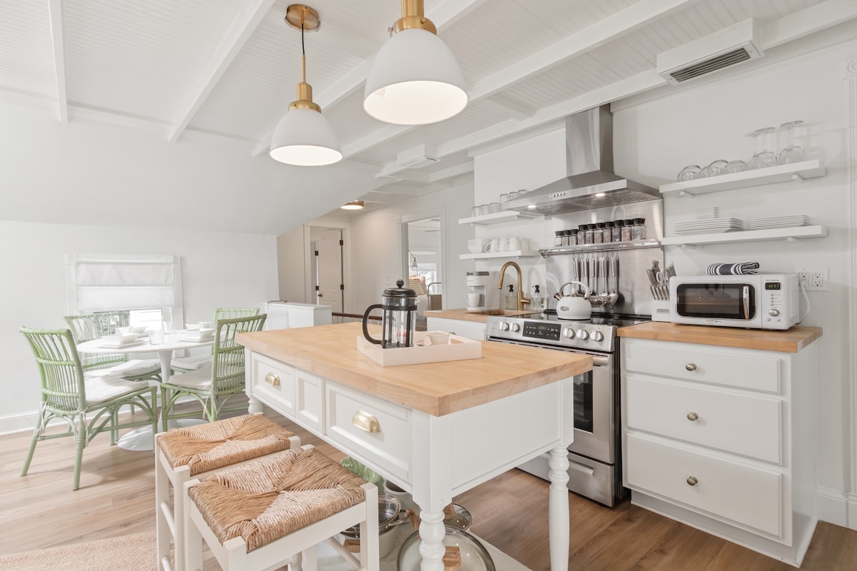 A well-equipped kitchen showcases modern appliances and a spacious center island with stools. White cabinetry and light fixtures complement the bright atmosphere. A dining area with green chairs is visible in the background, enhancing the open-concept layout.