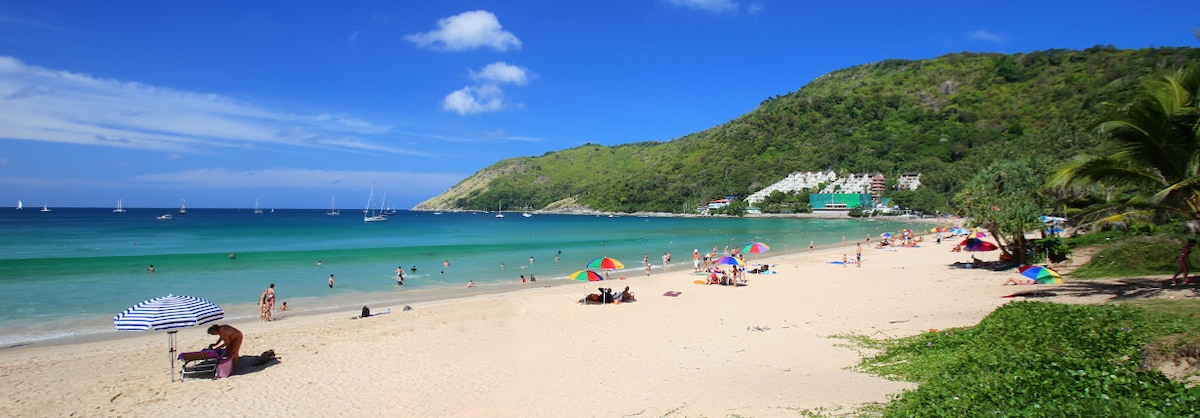 A scenic beach scene features soft sand and gentle waves in varying shades of blue and green. Sun umbrellas in vibrant colors dot the shoreline, and beachgoers are seen relaxing and enjoying the sun. Lush green hills rise in the background, providing a natural contrast.