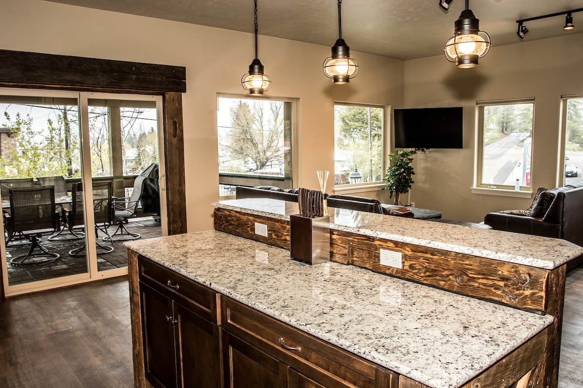 A modern kitchen area features a spacious granite countertop with wooden cabinetry. Large windows provide natural light and views of the exterior deck. Pendant lights hang from the ceiling, and a flat-screen TV is mounted on the wall nearby, enhancing the open and airy feel of the space.
