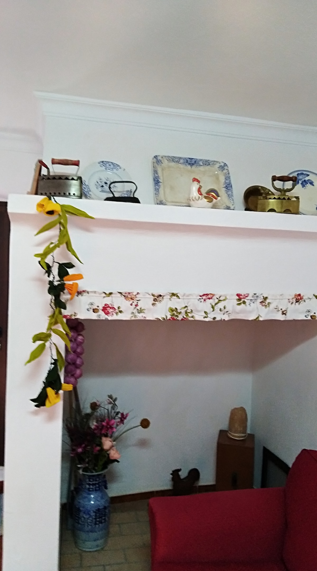 A decorative shelf holds vintage items such as old irons and ceramics, adding charm to the room. A floral-patterned curtain gently drapes below the shelf. A colorful floral arrangement is positioned in a vase nearby, enhancing the welcoming atmosphere of the space.