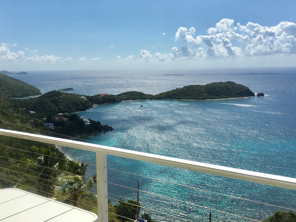 A panoramic view of the coastline showcases various islands surrounded by turquoise waters. The sky is bright with scattered clouds, and the landscape includes lush greenery along the shore, creating a serene and inviting atmosphere.