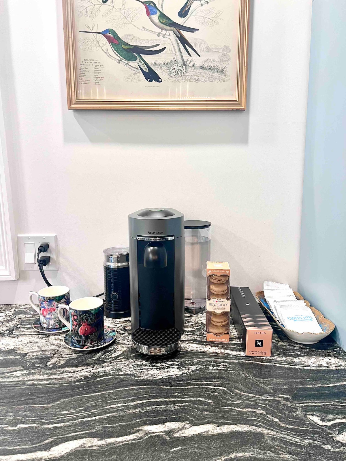 A coffee station is presented on a patterned granite countertop. A sleek coffee machine is positioned beside a glass carafe, with a selection of coffee and tea pods neatly arranged in a small tray. Two mugs with colorful designs rest on decorative coasters.
