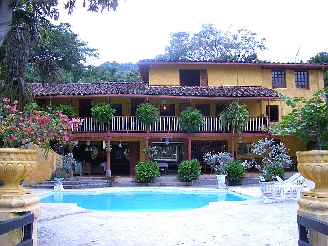 A two-story rustic ranch house is framed by lush greenery, featuring large windows and a balcony adorned with potted plants. A crystal-clear swimming pool sits in the foreground, surrounded by terracotta planters filled with vibrant flowers, creating a serene outdoor space.