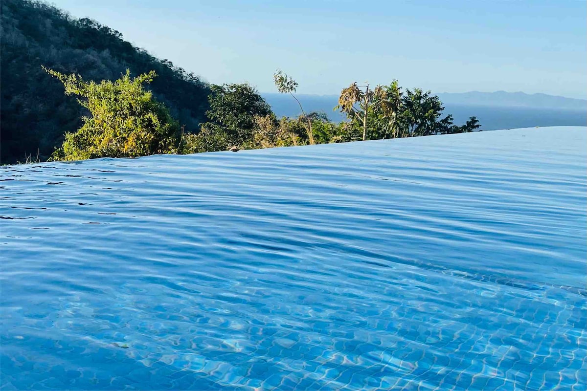 An infinity pool stretches out towards the horizon, blending seamlessly into the sky. Lush greenery surrounds the pool area, offering a serene backdrop of nature and distant mountains. The gentle ripples on the water reflect the calmness of the environment.