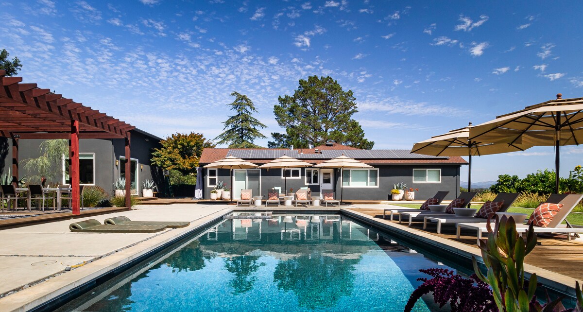 A large solar-heated pool reflects the surrounding sky, complemented by a wooden deck furnished with lounge chairs and umbrellas. The modern guest house is visible in the background, set among landscaped grounds featuring trees and plants.