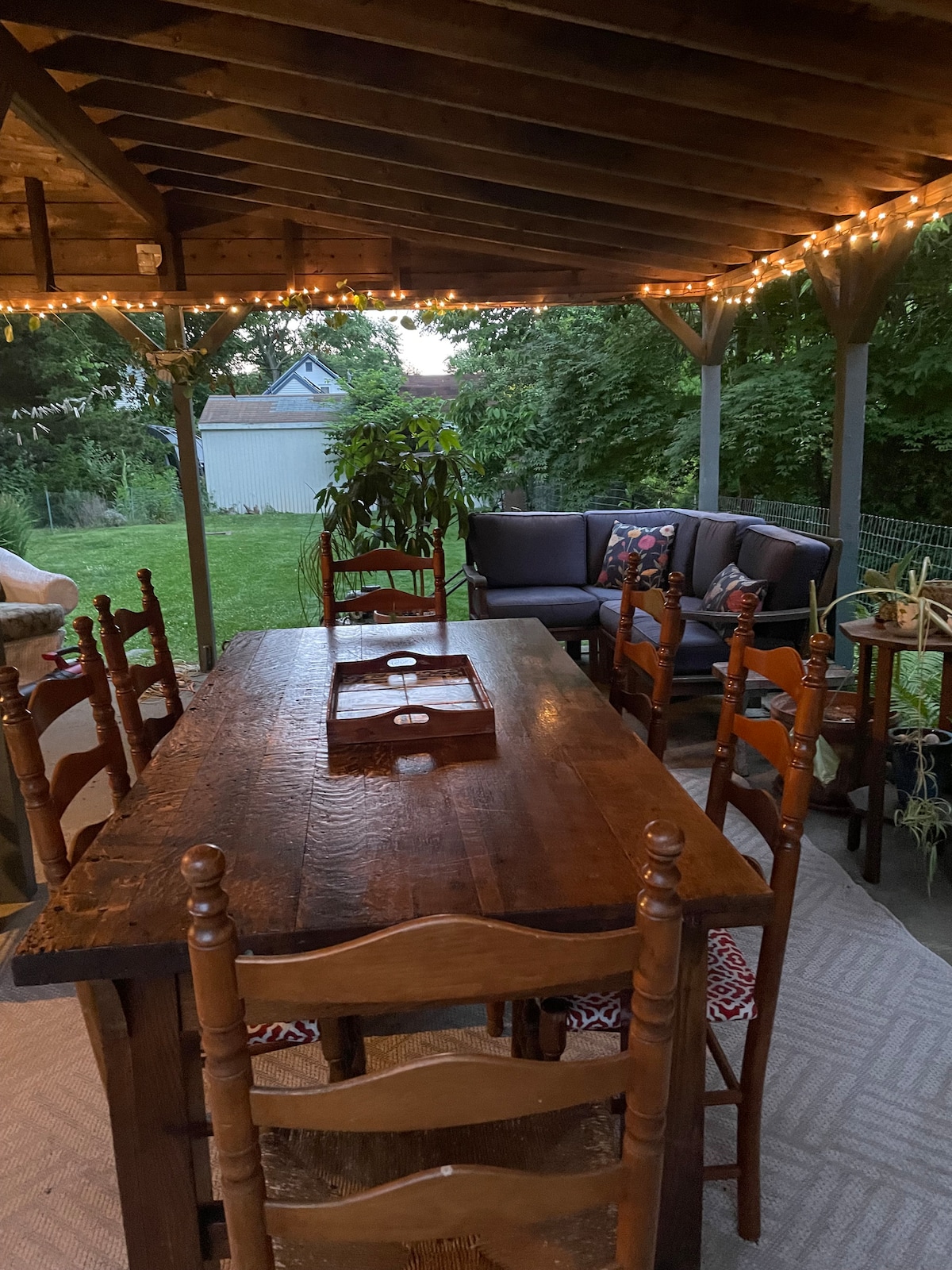 An outdoor patio area features a rustic wooden dining table surrounded by six chairs, partially illuminated by string lights overhead. A comfortable seating arrangement is positioned nearby, accompanied by potted plants. Lush greenery is visible in the background, creating a serene setting.