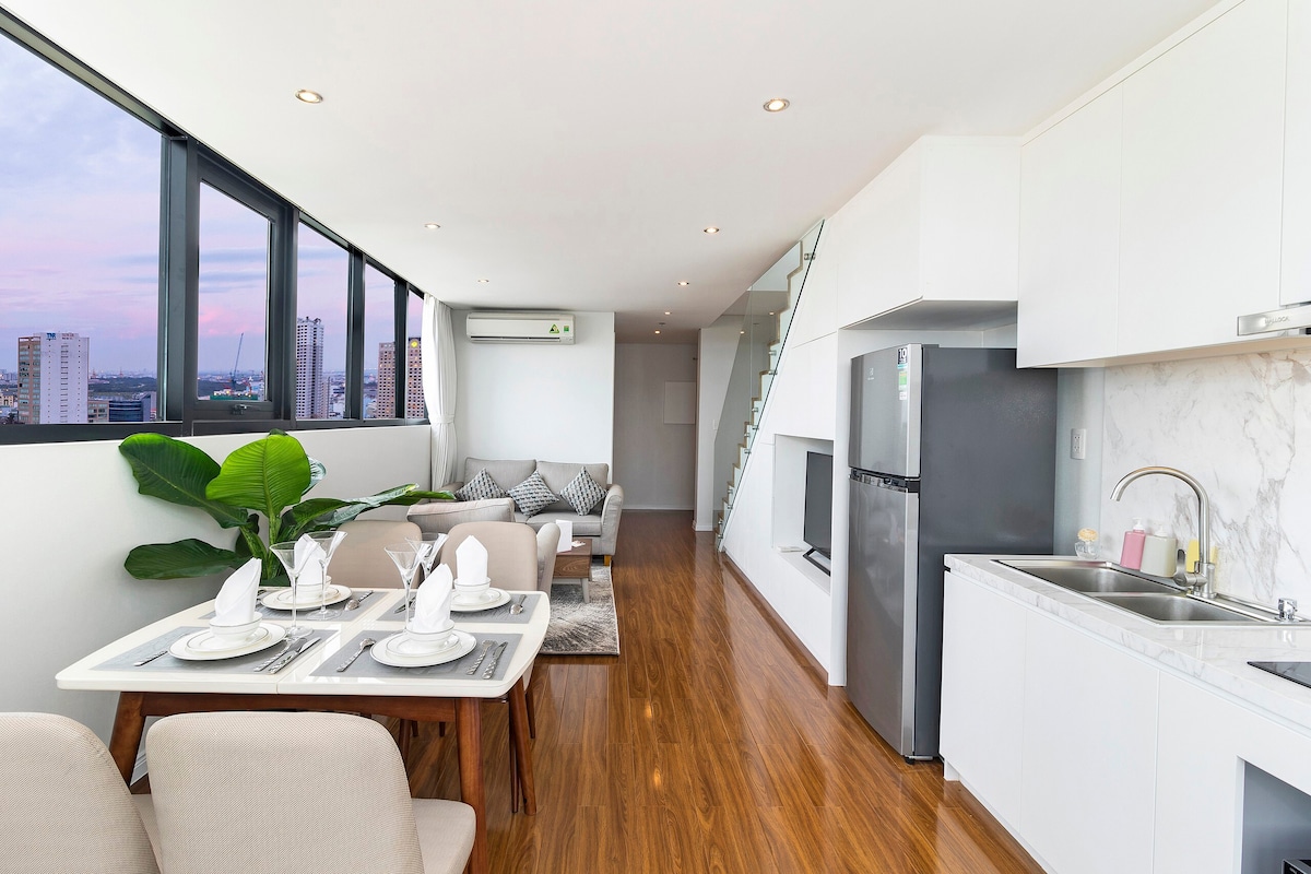 A spacious living area showcases a modern dining table set for four, with neatly arranged tableware. A comfortable seating area includes a sofa arranged with decorative pillows. Large windows provide natural light and offer a view of the city, enhancing the room's airy feel.