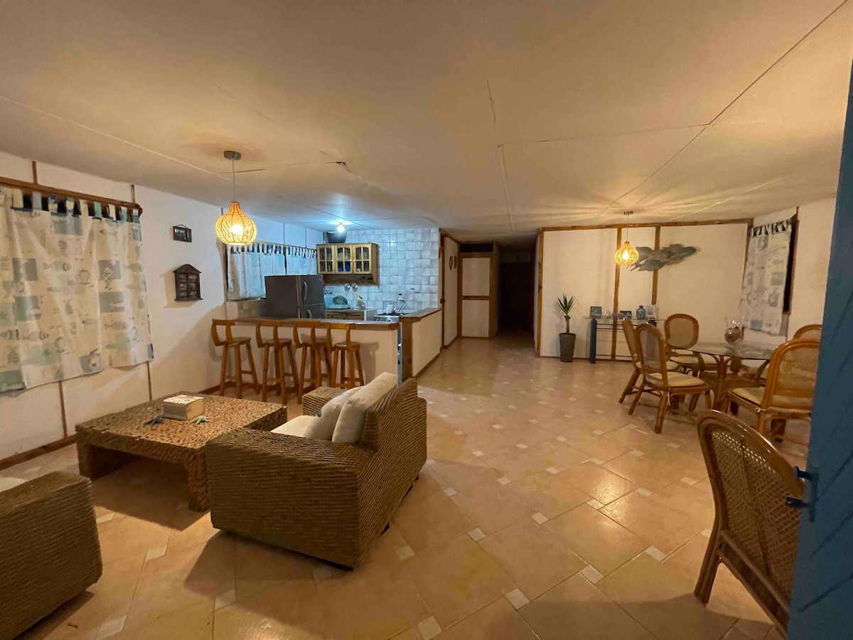 An open living area is displayed, featuring a seating arrangement made of natural fibers. A dining table is visible, surrounded by rattan chairs. The kitchen area includes wooden cabinetry and modern appliances, while a neutral-toned tiled floor complements the warm lighting.