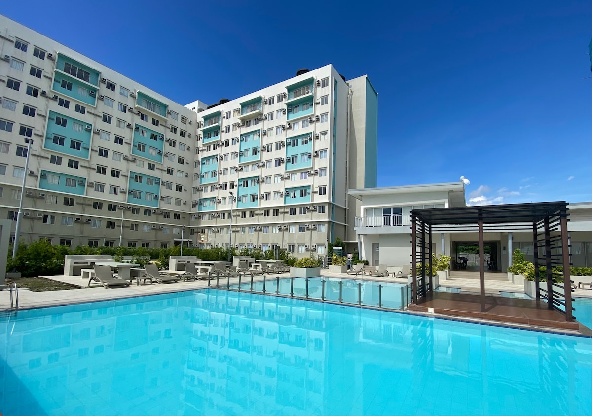 A spacious outdoor pool is framed by a well-maintained deck and lounge area. Surrounding buildings are visible with a modern design, including balconies. Lush greenery adds a refreshing touch, with clear blue skies above enhancing the inviting atmosphere of the community space.