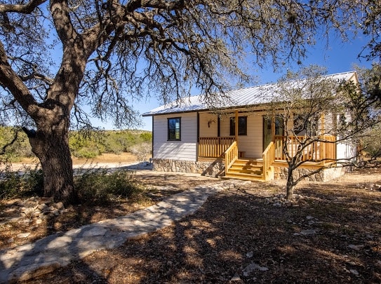 The charming farmhouse features a bright exterior with a welcoming front porch. A path lined with stones leads to the entrance, surrounded by natural vegetation and mature trees. The landscape includes rolling hills in the background under a clear blue sky.