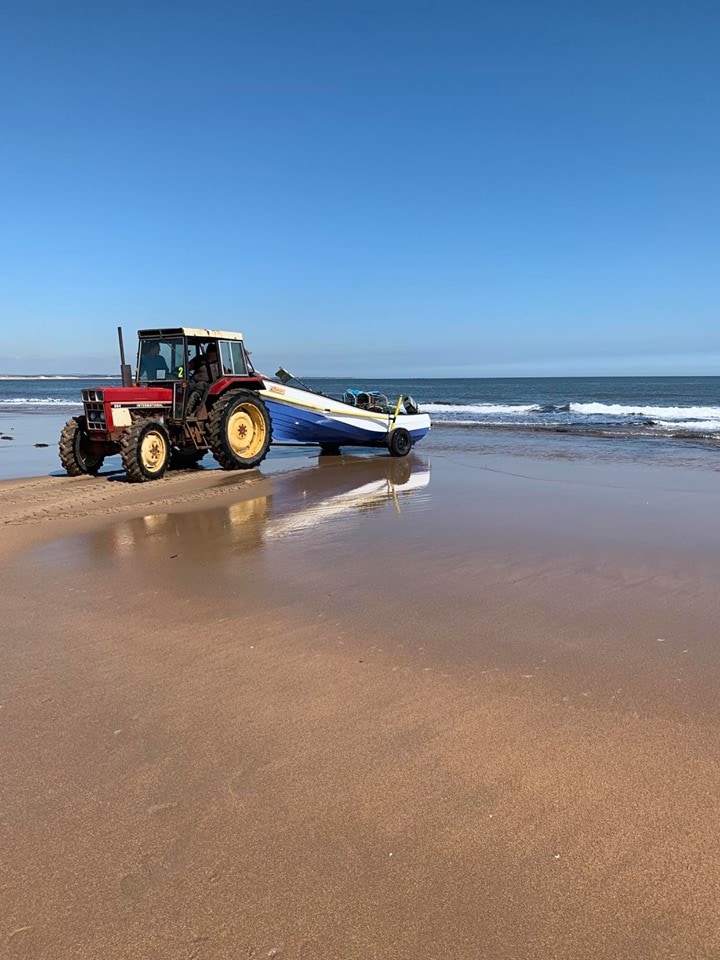 A tractor is positioned at the water's edge on a sandy beach, towing a boat towards the ocean. The clear blue sky contrasts with the gentle waves lapping at the shore, creating a serene coastal scene.