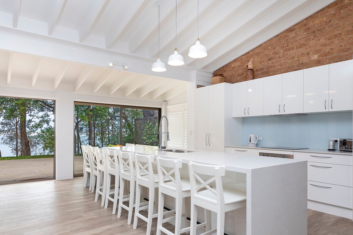 A spacious kitchen features a large white island counter with ample seating. Bright pendant lights hang above, complementing the white cabinetry. Large glass doors provide views of the surrounding trees and lake, enhancing the connection between indoor and outdoor spaces.