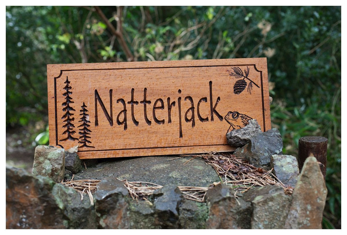 A wooden sign displaying the name 'Natterjack' is affixed to a stone wall. The sign features carved illustrations of trees and a small frog, with natural elements like pine needles scattered around it, blending harmoniously with the surrounding greenery.