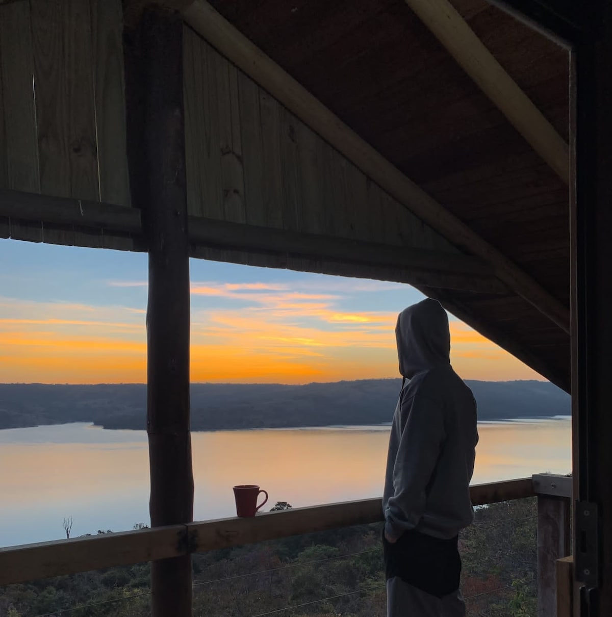 A person stands on a balcony framed by wooden posts, gazing at a serene lake at sunrise. Soft hues of orange and blue are reflected in the water, creating a tranquil view. A cup rests on the railing, suggesting a moment of relaxation.